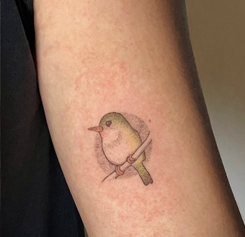 Small dotwork bird on a branch tattoo in soft green and gold on an upper arm